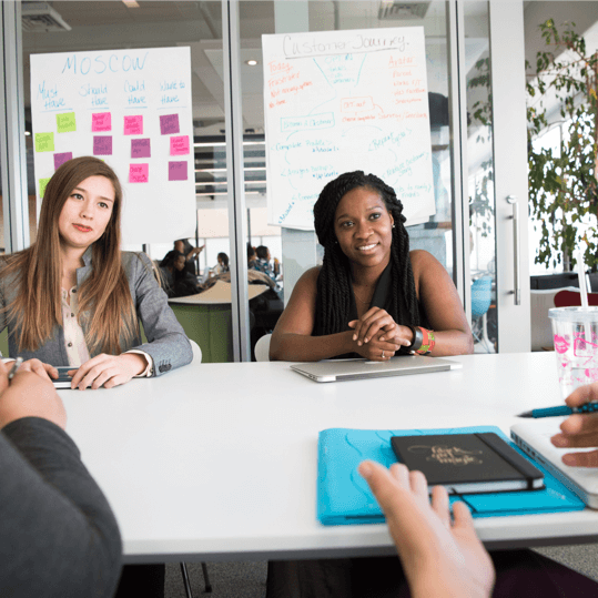 Two professionals sitting at a meeting table in a modern office, discussing plans with notes and a laptop visible, representing accessible workplace communication and client focused communication support.