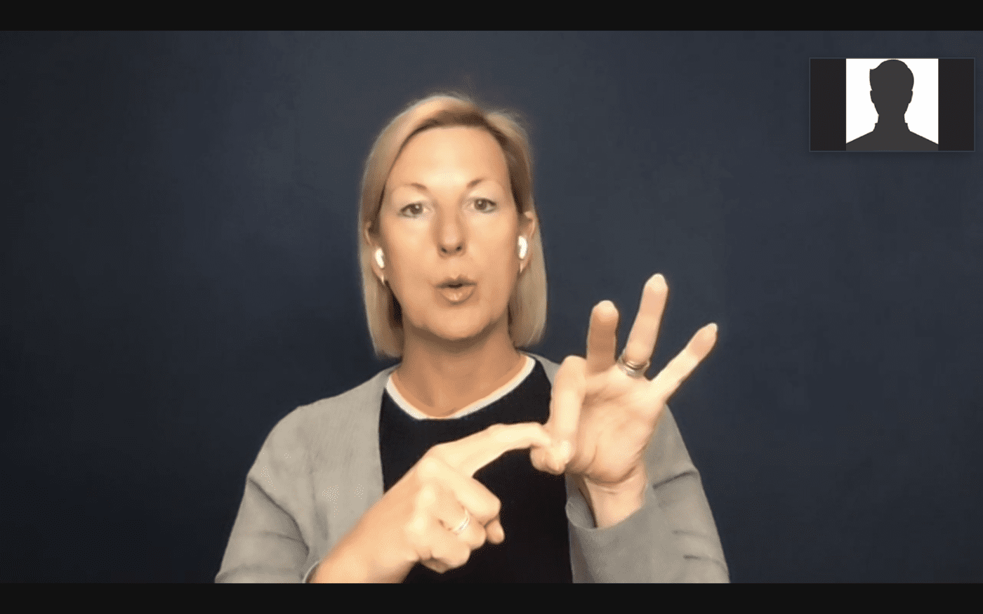 A woman with short blonde hair is signing in British Sign Language during an online video call. She is wearing earphones and a grey cardigan over a black top, and is pointing to her left hand while making a sign. The screen shows a small participant window in the top right corner.