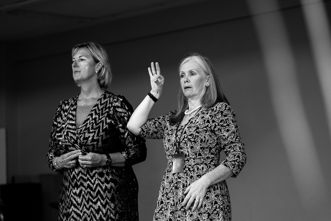 Two women stand on a stage. One is Lipspeaking. The other is signing in British Sign Language. Both are wearing patterned dresses and facing forward. The image is in black and white.