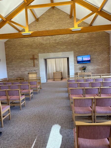 The inside of a crematorium. The chairs are all empty and there is a cross visible on the wall.
