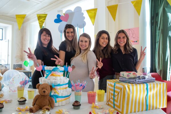 A group of women are posing for a picture, stood behind a table filled with presents and a cake. One of the women are visibly pregnant.