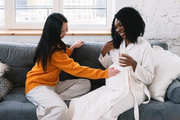 Two women are sat on a soft. One woman is reaching over to place her hand on another woman's baby bump.