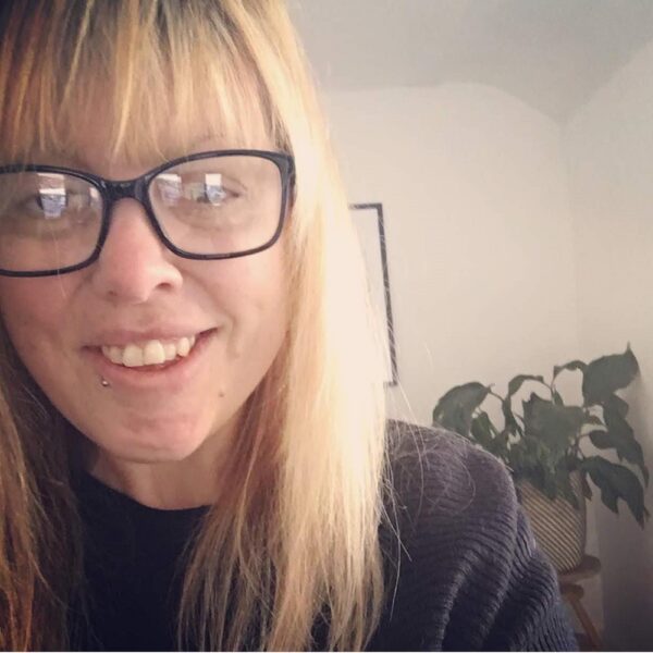 A profile photo of a woman with long blonde hair and black glasses. She is wearing a black jumper. In the background you can see the edge of a photo frame on the wall and a plant.
