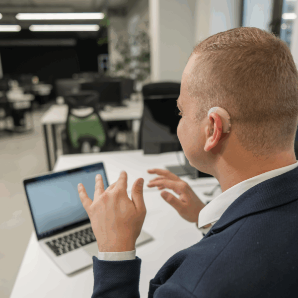 A deaf man wearing a hearing aid sits at a desk in an office, communicating using sign language during a video call on his laptop. The office space around him is modern and mostly empty.