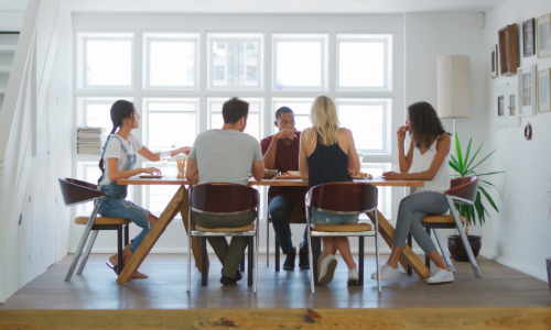 Five people sit around a wooden dining table in a bright room with large windows behind them. They are mid-conversation and eating together.