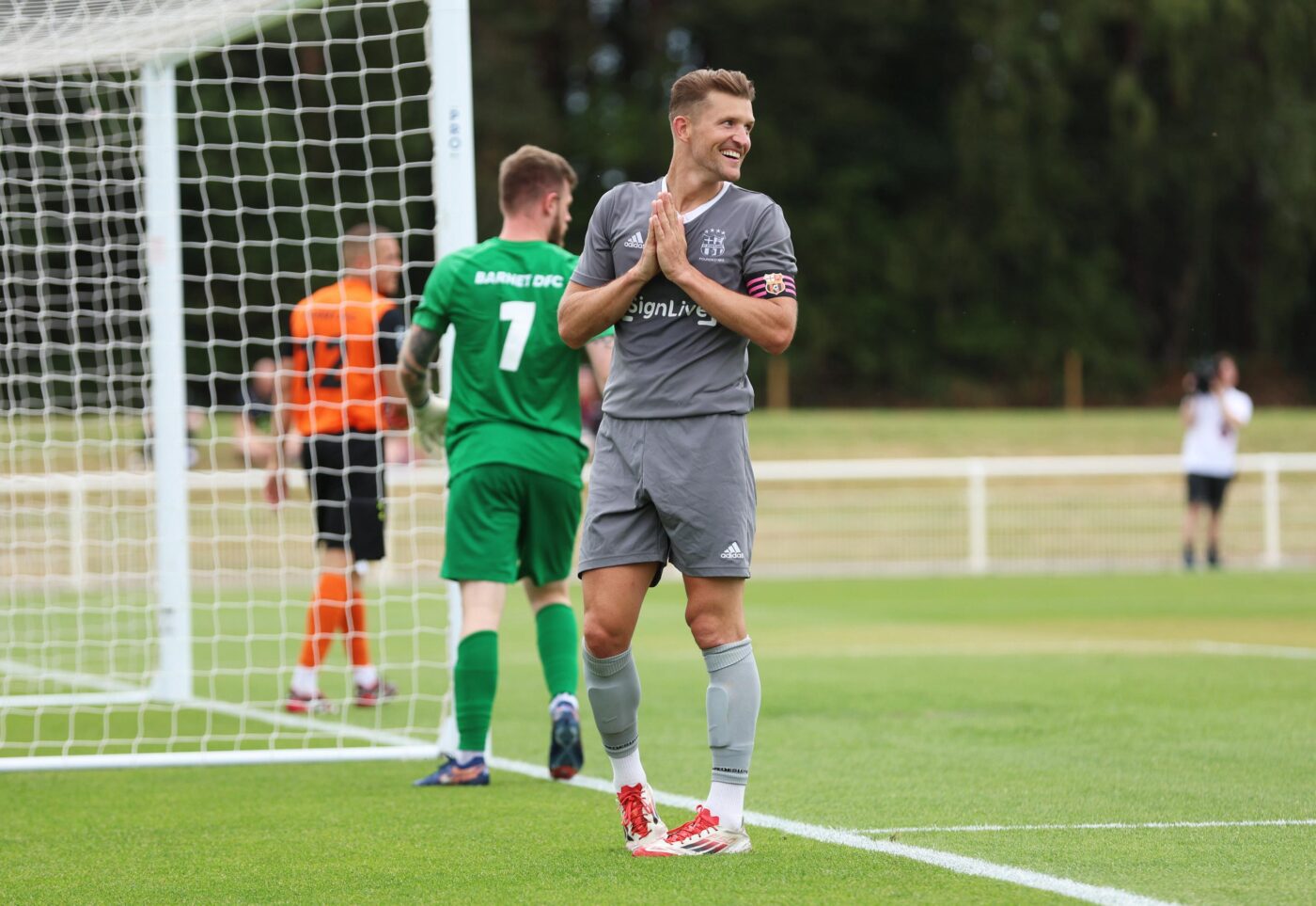 Jamie Clarke, a deaf footballer, smiling and clapping on the pitch during a match, wearing a grey kit with a pink captain’s armband.