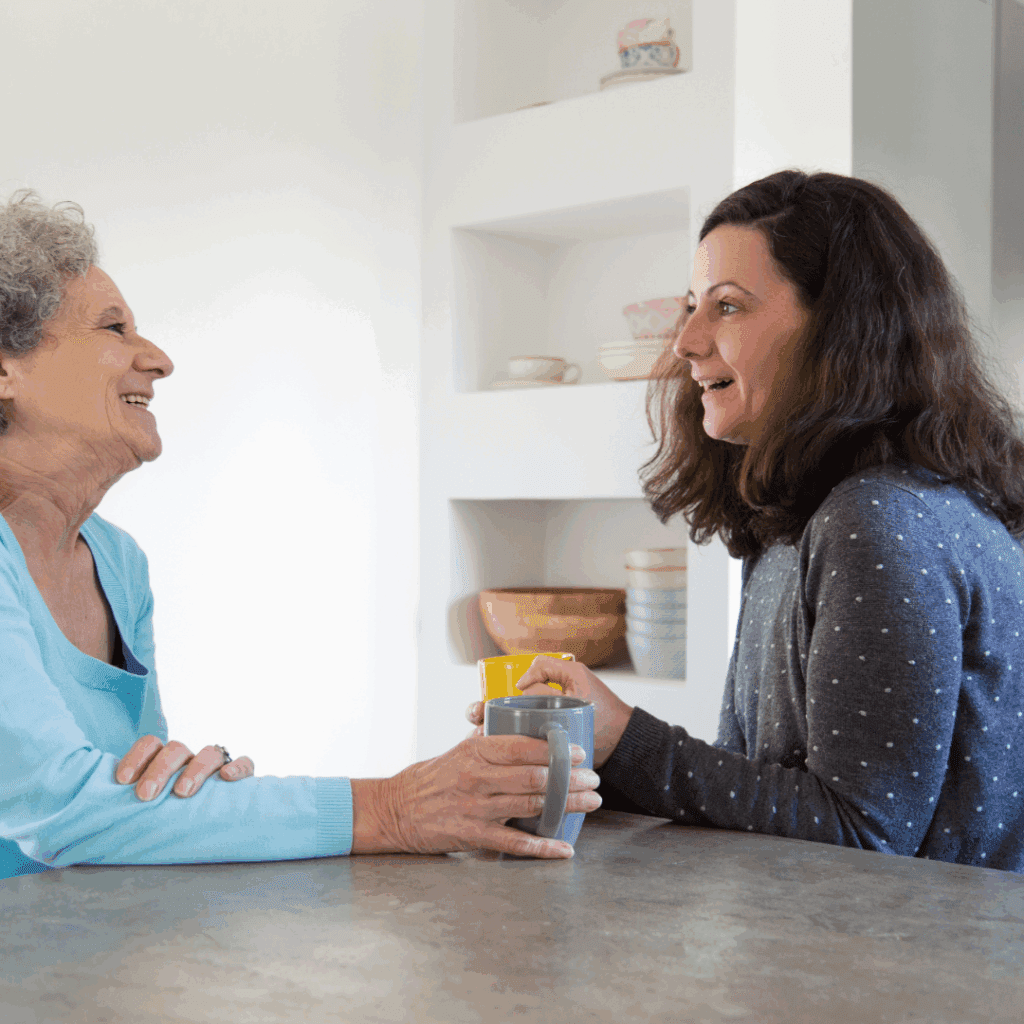 Two women sit at a kitchen table, smiling and talking while holding mugs, facing each other closely.